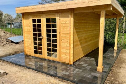 Wooden garden shed with glass-paneled French doors on a tiled patio, outdoors on a sunny day.