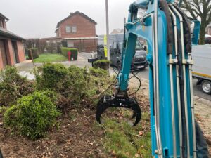 Blue hydraulic excavator arm extended over a suburban street with a grey van and brick houses in the background, logs and shrubs in the foreground.