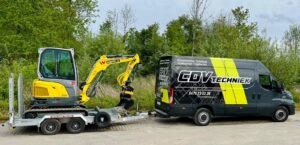 Yellow mini excavator on a silver trailer hitched to a dark gray CDV Techniek van, parked in a green, tree-lined area on a paved lot.
